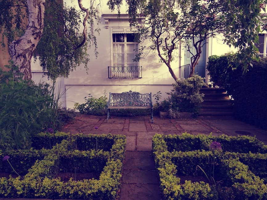 A small, well-maintained garden attached to a white residential building, featuring a paved patio area with red bricks, a decorative metal garden bench in front of the house, and neatly trimmed low bo